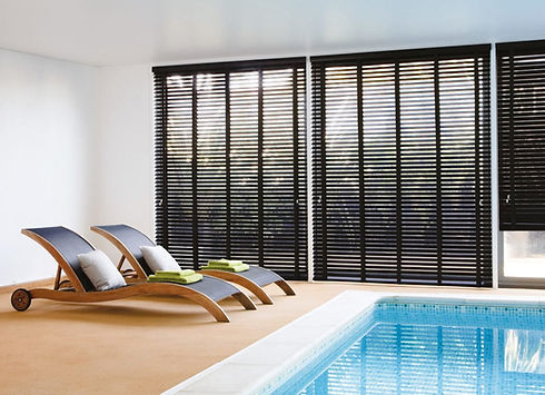 Modern indoor pool area with two wooden lounge chairs on tan flooring, green towels, cushions, and black vertical blinds creating a tranquil ambiance