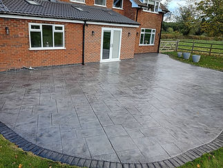 A polished, stamped concrete patio with a tile-like pattern extends from a brick house with white-framed windows and doors, bordered by greenery