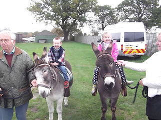 Children joyfully ride donkeys led by adults on a grassy field