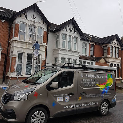 Painter on a ladder decorating the white bay window frames of a house exterior behind a company van