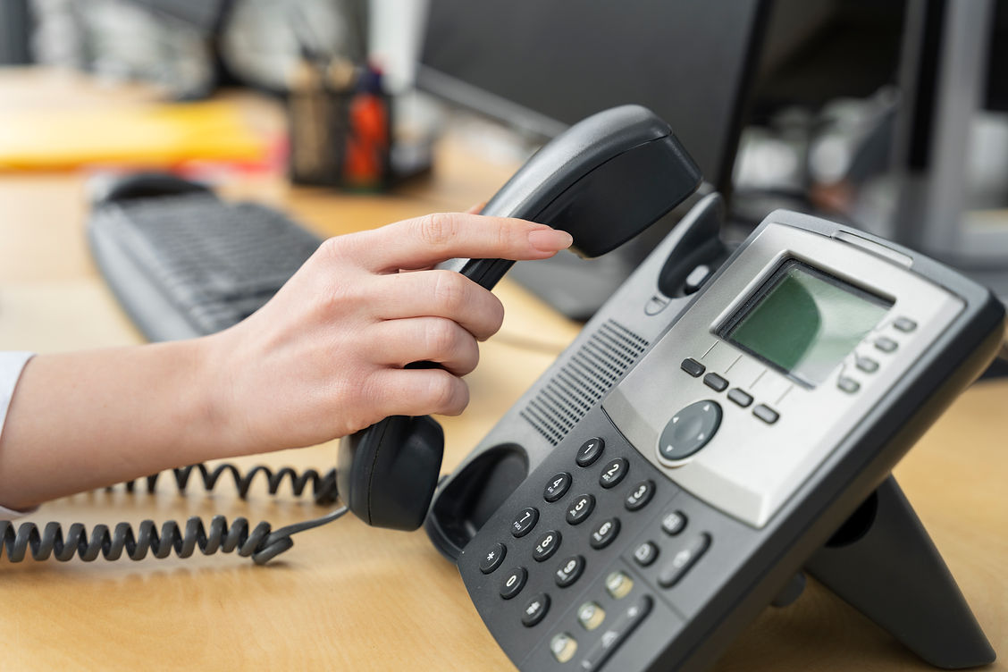 A hand lifts the receiver of a black office phone on a wooden desk, surrounded by computer equipment, conveying professionalism and communication