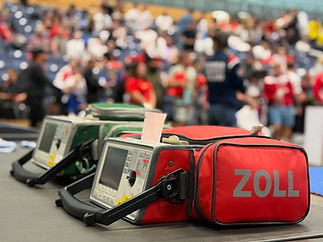 Three defibrillators in protective cases, foreground; two green, one red labeled "ZOLL." Blurred crowd in background, suggesting a public event setting