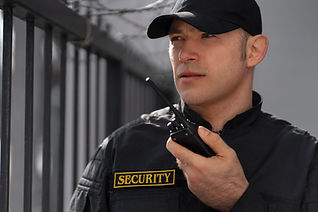 Security guard in a black uniform and cap holds a walkie-talkie, standing alert by a metal fence with barbed wire, conveying vigilance