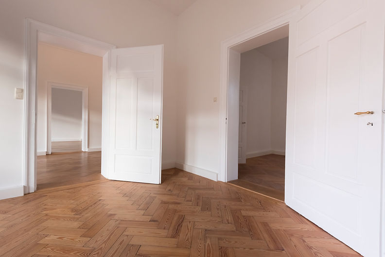 Spacious, empty room with open white doors, light wood herringbone floor, and soft white walls