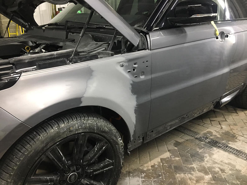 A silver car in a workshop with an open hood and partially sanded front fender, revealing a matte finish