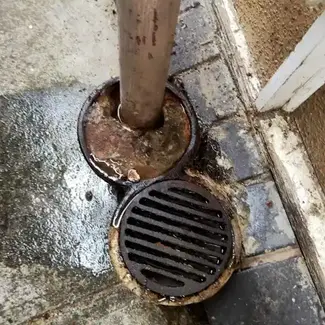 A rusty drainpipe overflows next to a wet metal grate on a damp sidewalk