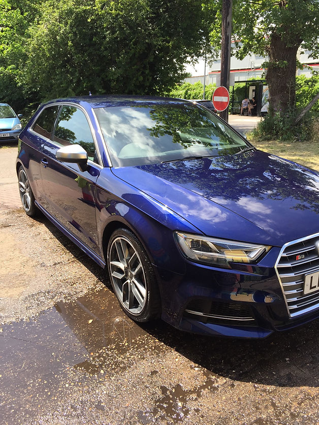 Front three-quarter view of a clean, deep metallic blue Audi S3 hatchback parked on a sunny day next to a puddle, reflecting green trees in its paint and windshield