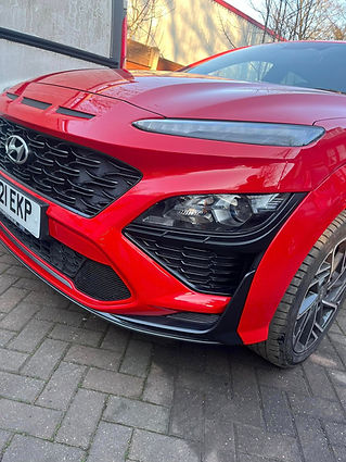 Red sports car showing a sleek front design with a prominent grille, sharp headlights, and a visible license plate, parked on a brick driveway