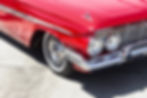 Close-up of a vintage red car's front corner, featuring shiny chrome accents, round headlights, and whitewall tires