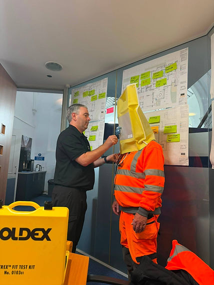 Trainer conducting a face-fit test on a worker wearing a yellow testing hood and orange high-visibility clothing