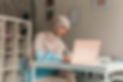A woman in a beige hijab and blue shirt writes in a notebook at a white table with a laptop