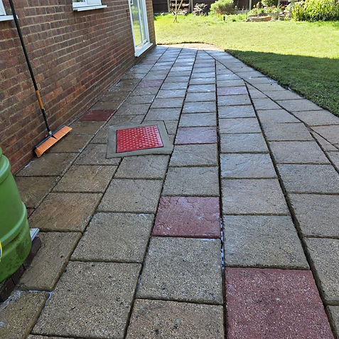 Clean paved stone patio and pathway with alternating grey and reddish accent slabs running along a brick house wall toward a grassy lawn