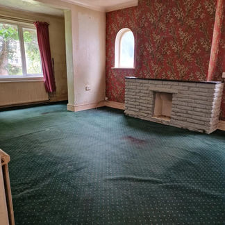 An empty living room with a green carpet, a fireplace, patterned red wallpaper, and a small arched window
