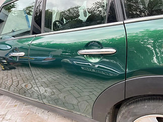 Green car door with a sleek design and chrome handle, reflecting surrounding trees and pavement patterns, conveying a polished and reflective appearance