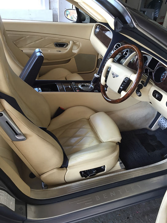 Interior view of a luxury Bentley convertible featuring cream-colored quilted leather seats, wood-trimmed steering wheel, and detailed dashboard controls