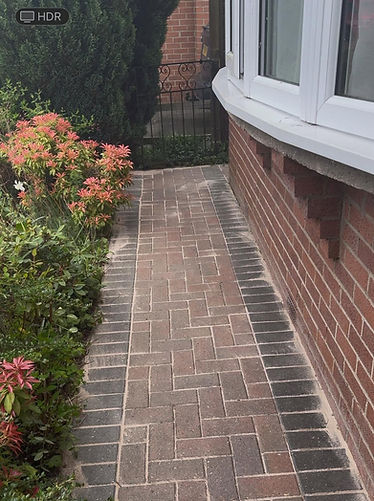 Block-paved garden pathway with a dark border, running alongside a red brick house wall with a bay window and leafy shrubs