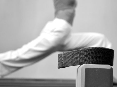 Girl at the back in white pants doing warrior 1 pose, in front a yoga block and strap.
