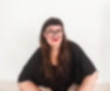 Portrait einer entspannt sitzenden Person mit Brille, roten Lippen und langem Haar vor minimalistischem, hellem Hintergrund.