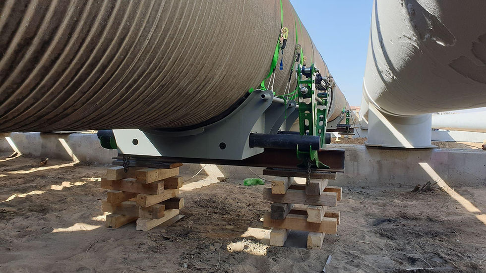 Large pipes supported by green braces rest on wooden blocks in a sandy construction site. Clear blue sky in the background.