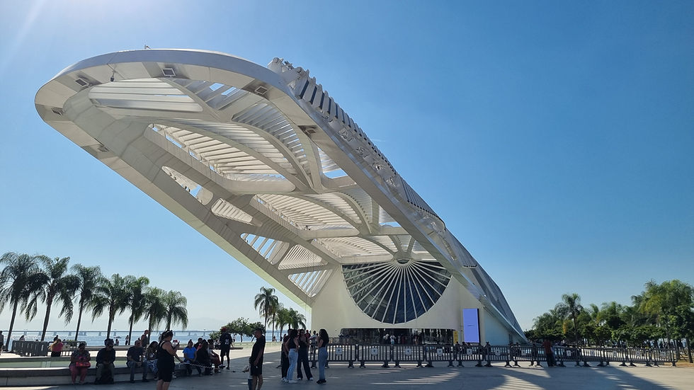 People gather under a futuristic white waterfront structure, with palm trees and a clear blue sky on a sunny day.