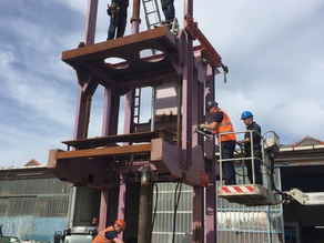 Workers inspect and assemble a tall purple industrial press on a construction site, with a cloudy sky and a lift platform