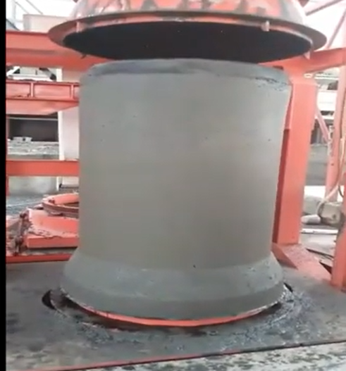 Gray cylindrical concrete pipe mould under a red concrete pipe machine press in a factory, with a heavy, work-in-progress look.
