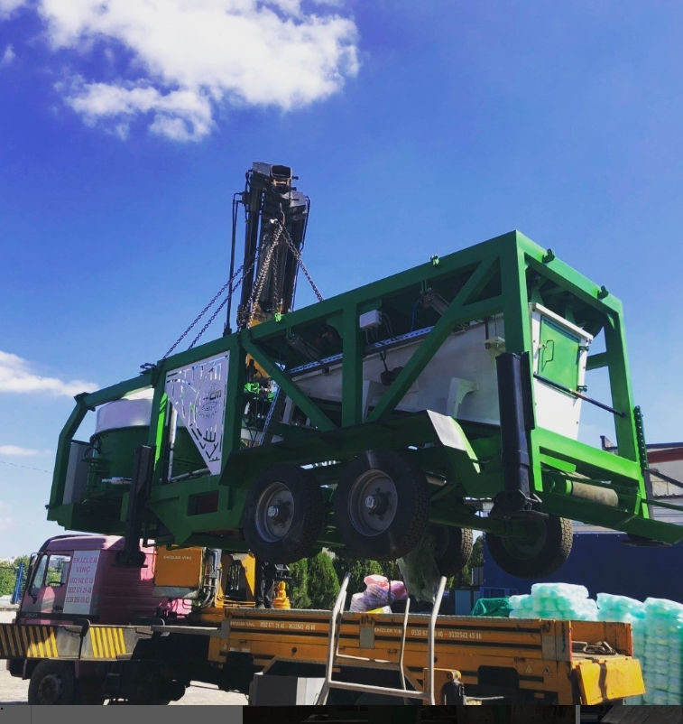 Green mobile concrete plant machine is hoisted by crane onto a flatbed truck under a blue sky with clouds.