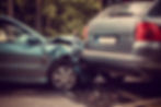Two cars in a rear-end collision on a forest-lined road, one silver and one teal, with visible damage and debris on the ground.