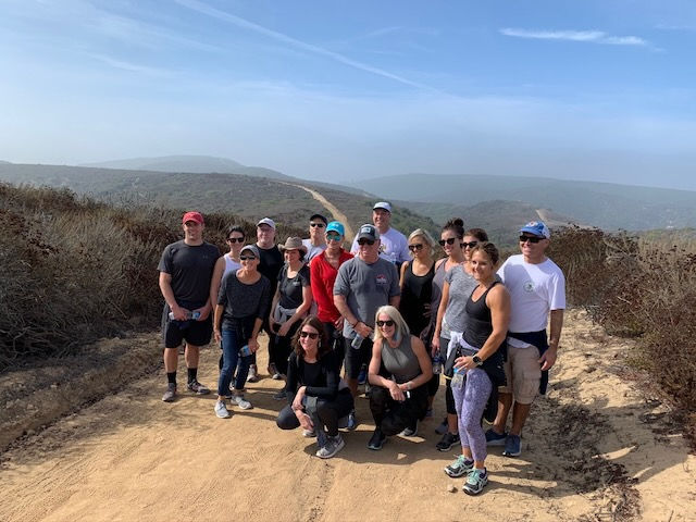 A group of 15 people pose on a sunny hiking trail with hills in the background. They're dressed in casual athletic wear, smiling and relaxed.