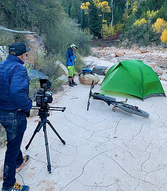 By a river with fall colored trees, Bhushan Thakkar behind the camera, in front Richie Schley sets up camp after a mountain bike ride.