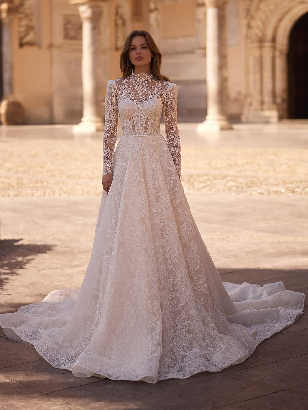 O vestido de noiva Aria Bride na Templo Fashion é uma fusão deslumbrante de elegância vintage e sofisticação moderna. Confeccionado inteiramente em delicada renda soutache, apresenta gola alta e mangas longas transparentes que conferem um toque de majestade. O corpete estruturado, com barbatanas internas, valoriza a silhueta ao mesmo tempo em que cria uma estética romântica e marcante. Um cinto fino com laço na cintura realça sutilmente as formas, adicionando um toque delicado e feminino. A peça flui para uma saia volumosa em corte evasê, onde o padrão da renda se mantém em perfeita continuidade, proporcionando um movimento suave e etéreo. Nas costas, o vestido desce em uma imponente cauda estilo capela, que irradia uma elegância atemporal. Combinado com um véu de acabamento em renda, o visual transmite uma majestade natural e sofisticada.
