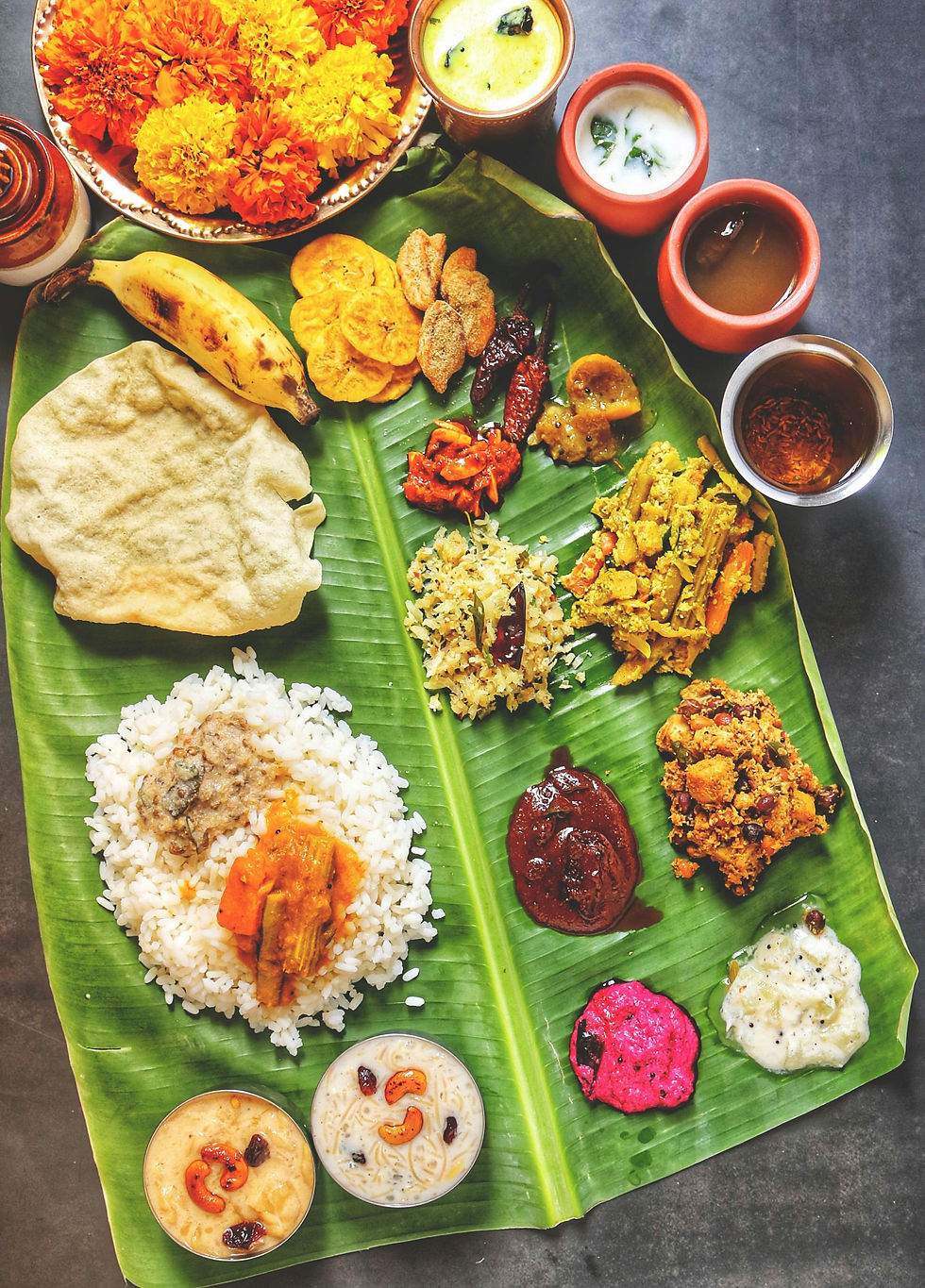 On a banana leaf, rice is in a small pile, surrounded by several curries and bowls of yoghurt and sauces. 