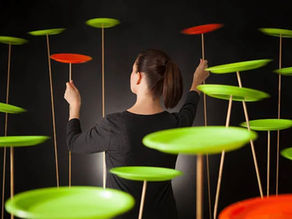 Woman in black, seen from behind, reaching toward green and orange spinning discs on sticks against a dark background.