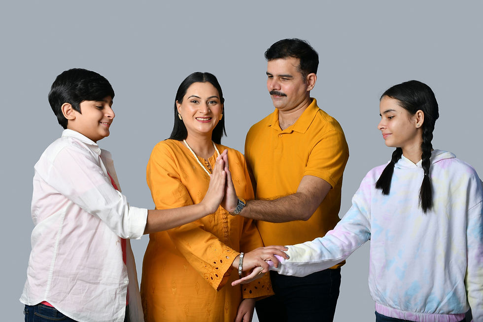 Smiling family of four poses against a gray backdrop, hands joined in the center; mother in orange, father in yellow, kids in white pastel tops