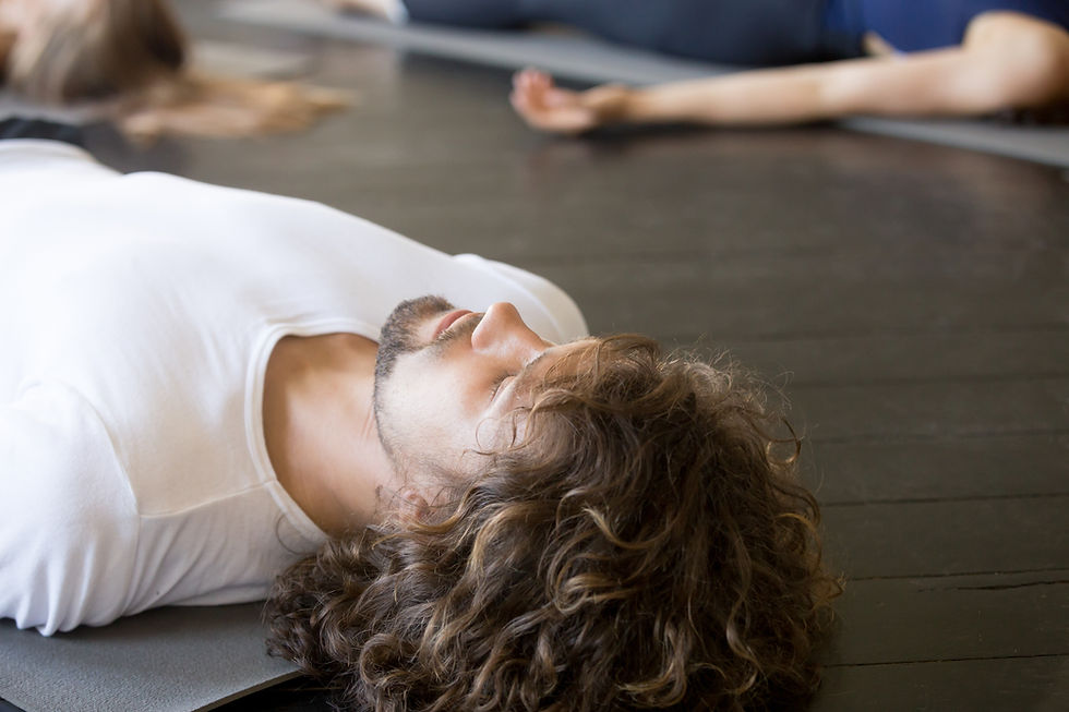 Man in a white shirt lies on a yoga mat during a group floor exercise, with blurred classmates in the background.