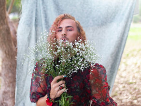 A man with a curly bob is wearing a rosy blouse and is holding a bouquet of baby's breath. Behind is a fabric, for what appears to be a photoshoot.