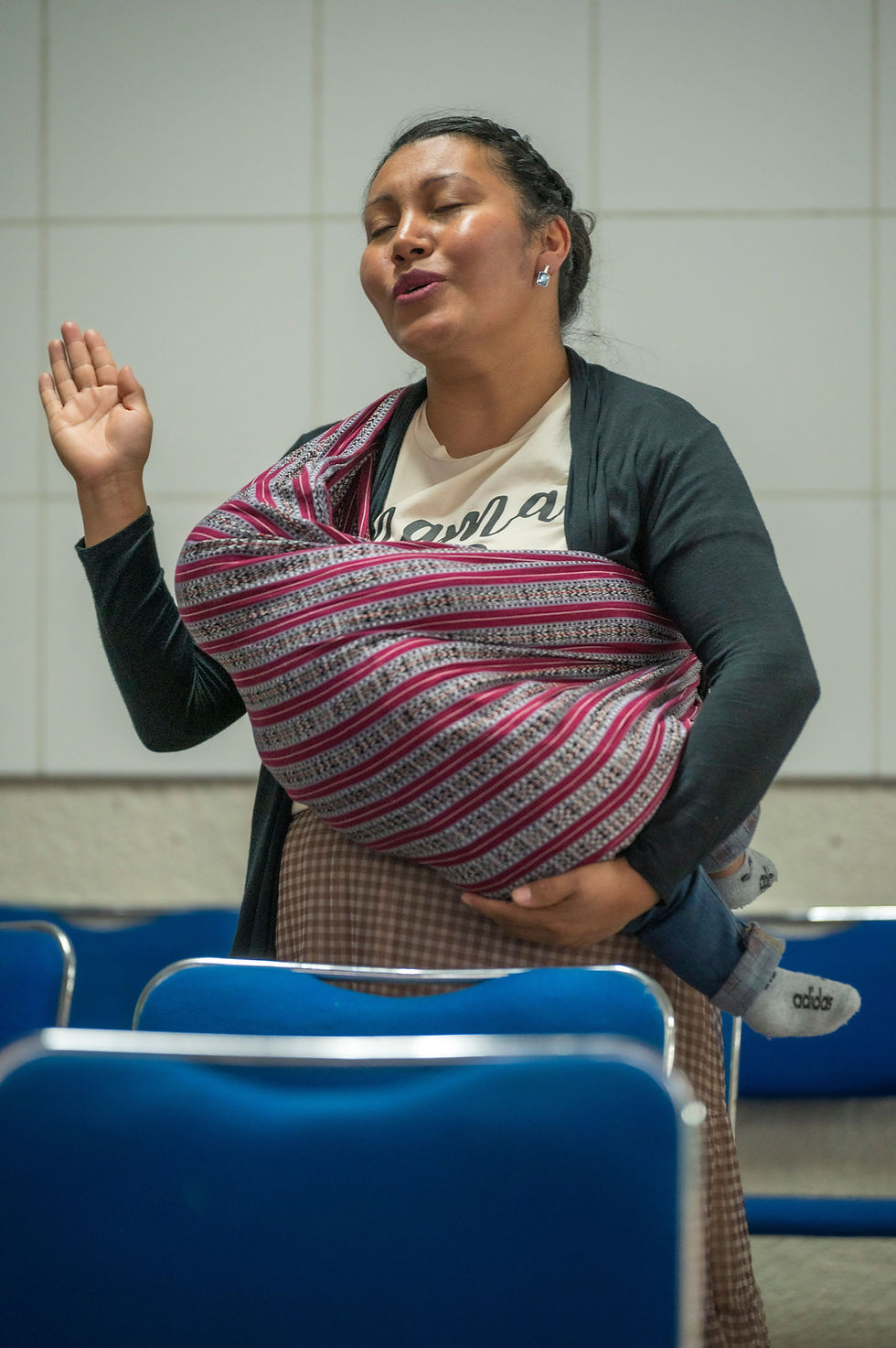 Woman with eyes closed raises a hand while carrying a baby in a striped sling, standing among blue chairs indoors.