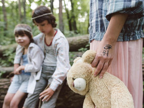 The image shows the lower half of a woman, with one hand holding a teddy bear. On a log next to her are seated two young boys. This is set in a forest. 
