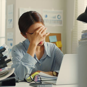A stressed woman, with her hand covering her face. She is sitting on a desk, covered with piles of paperwork and her laptop. 