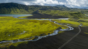 Aerial view of mossy green volcanic hills, black sand, winding river and road under a cloudy sky.