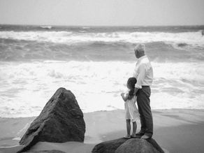 Black and white image of a grandfather and granddaughter standing together on rocks at the beach.