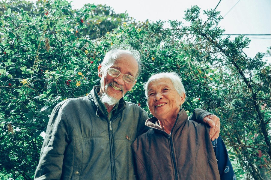 An elderly couple is smiling. The man has his arm around the woman, and behind a green bush mixed with coloured leaves. 