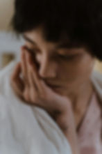 Close-up of a person with short dark hair, eyes closed, resting a hand on their face, wearing a white shirt in soft light