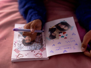 Hands write in a pink scrapbook with taped photos, heart stickers and a purple pen; handwritten note says I love you.