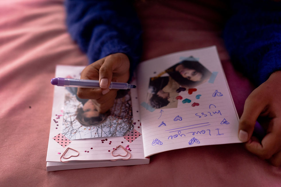 Hands write in a pink scrapbook with taped photos, heart stickers and a purple pen; handwritten note says I love you.