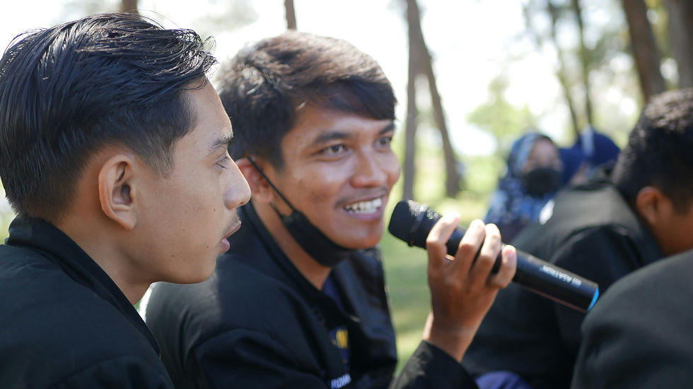 Two young men in black shirts outdoors; one smiles while speaking into a microphone, with trees and other people blurred behind.