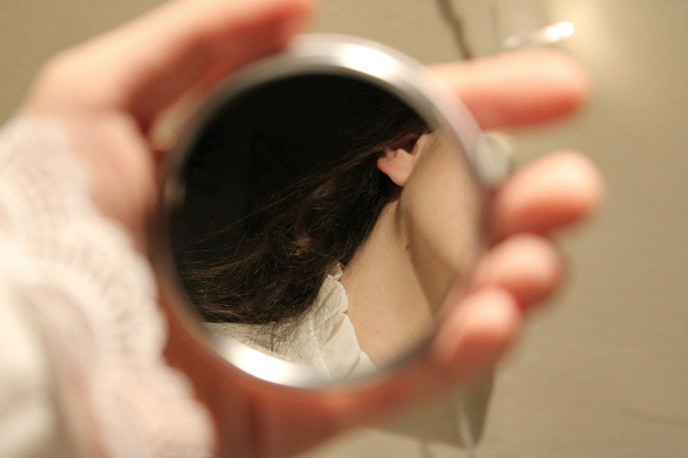 Hand holding a small mirror reflecting the back of a woman’s head and neck, with dark hair and a warm indoor blur.