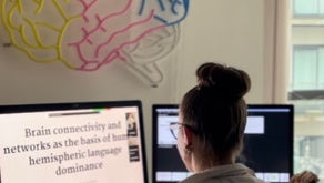 A woman wearing a white lab coat is standing in front of her desktop with her arms slightly raised. Above her desk is a multicoloured figure of a brain.
