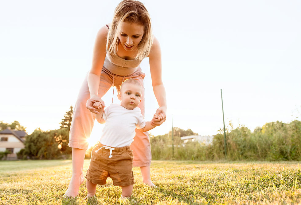 In a grassy field, a mother holds her baby's hands while the baby attempts to stand.