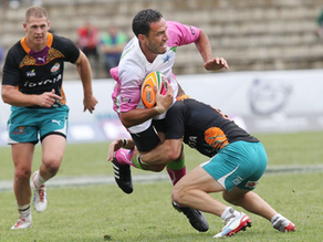 Rugby player in white and pink is tackled while carrying the ball, with a teammate running behind on the field.